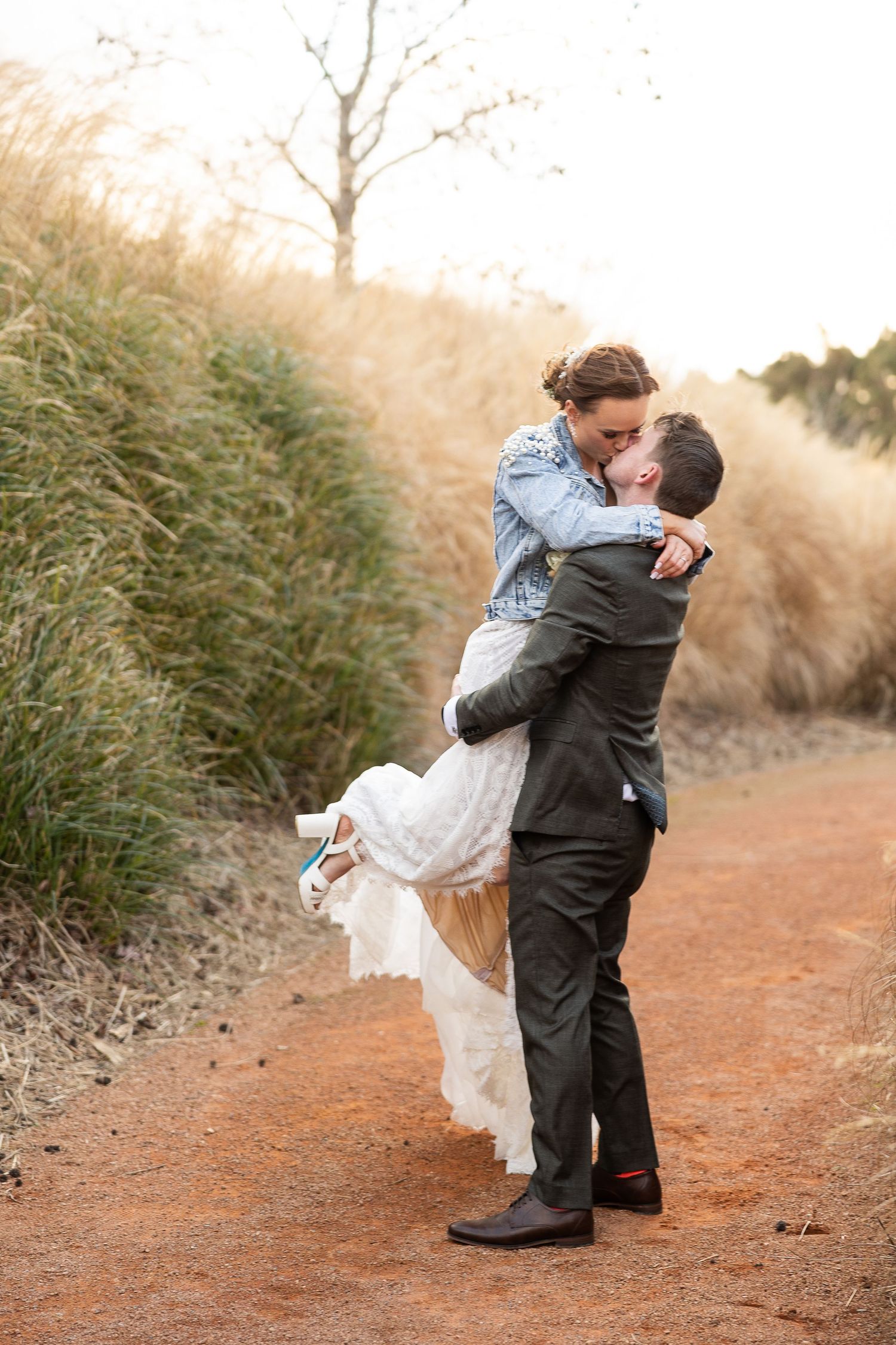 A romantic couple embraces on a rustic dirt path surrounded by tall grasses during an outdoor photo shoot.