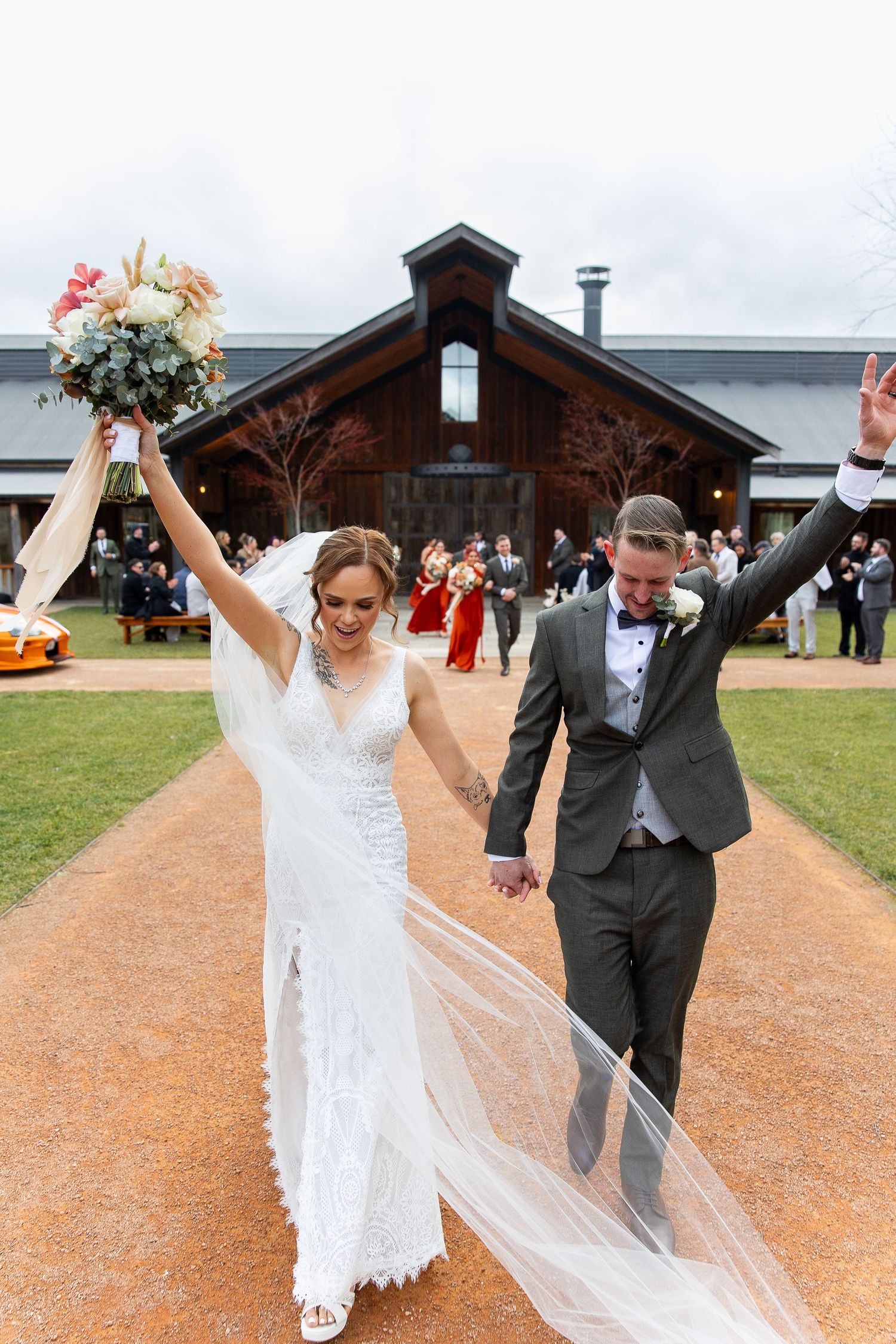 Joyful newlyweds celebrate as they walk on a gravel path with rustic wooden barn buildings in the background.