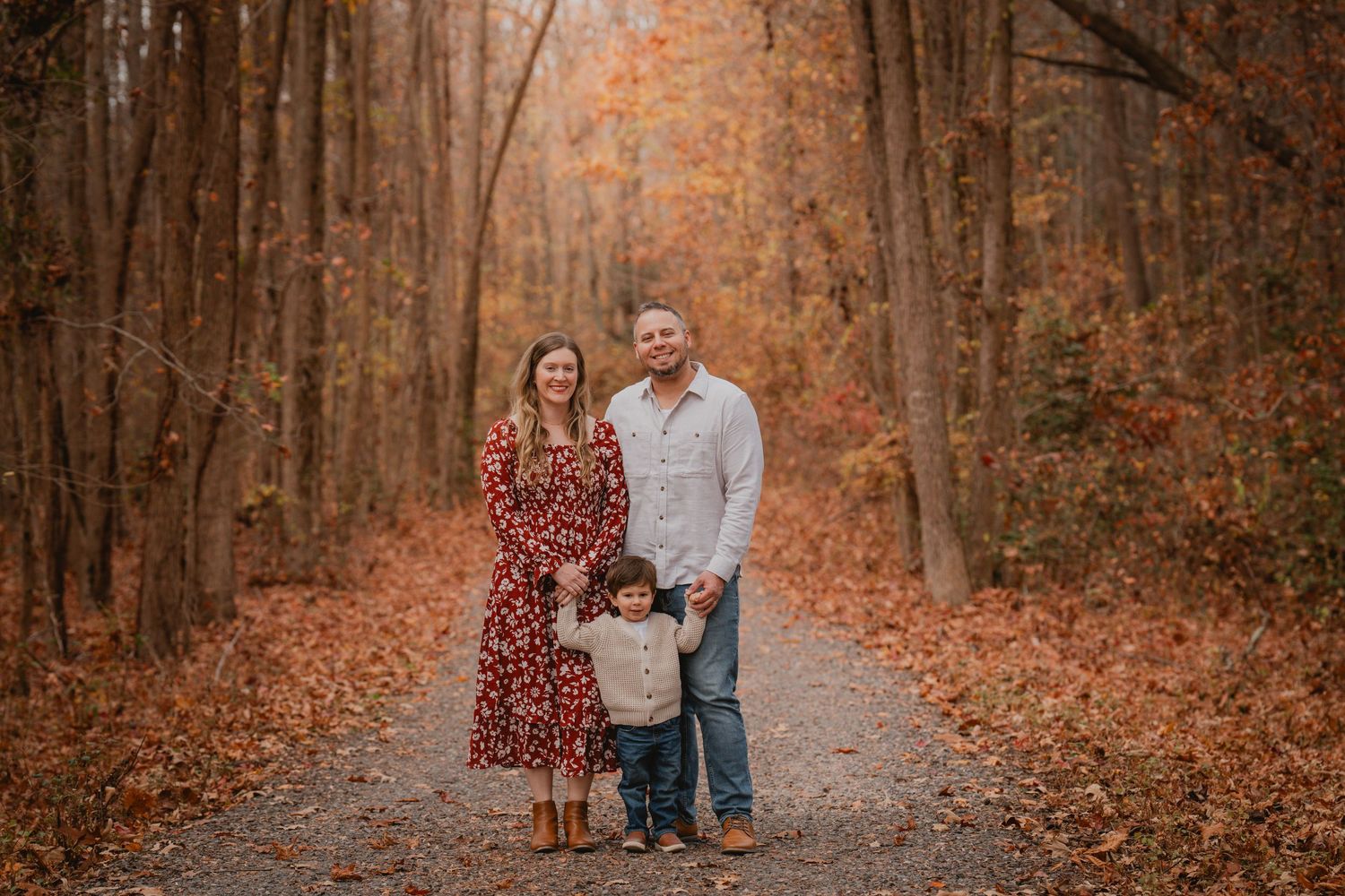 Family of three standing on a leaf-covered path surrounded by vibrant fall foliage.