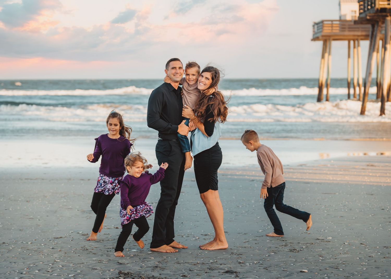 Joyful family of six playing together on a sandy beach at sunset near a pier.