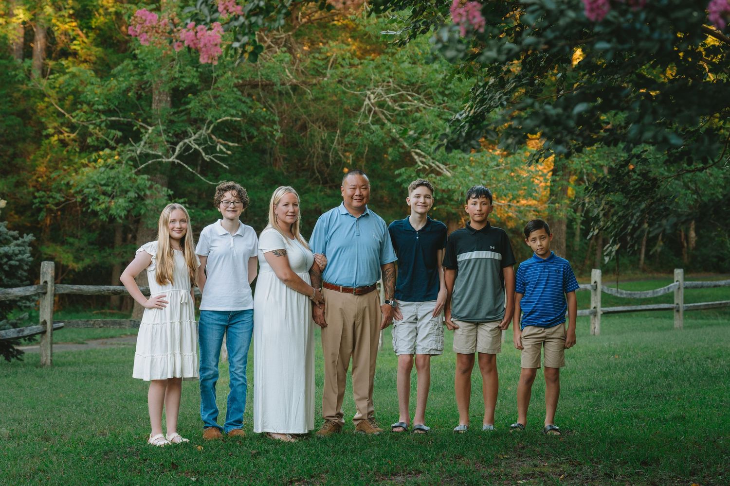 Family of seven smiling together outdoors in a lush green yard with colorful trees in the background.