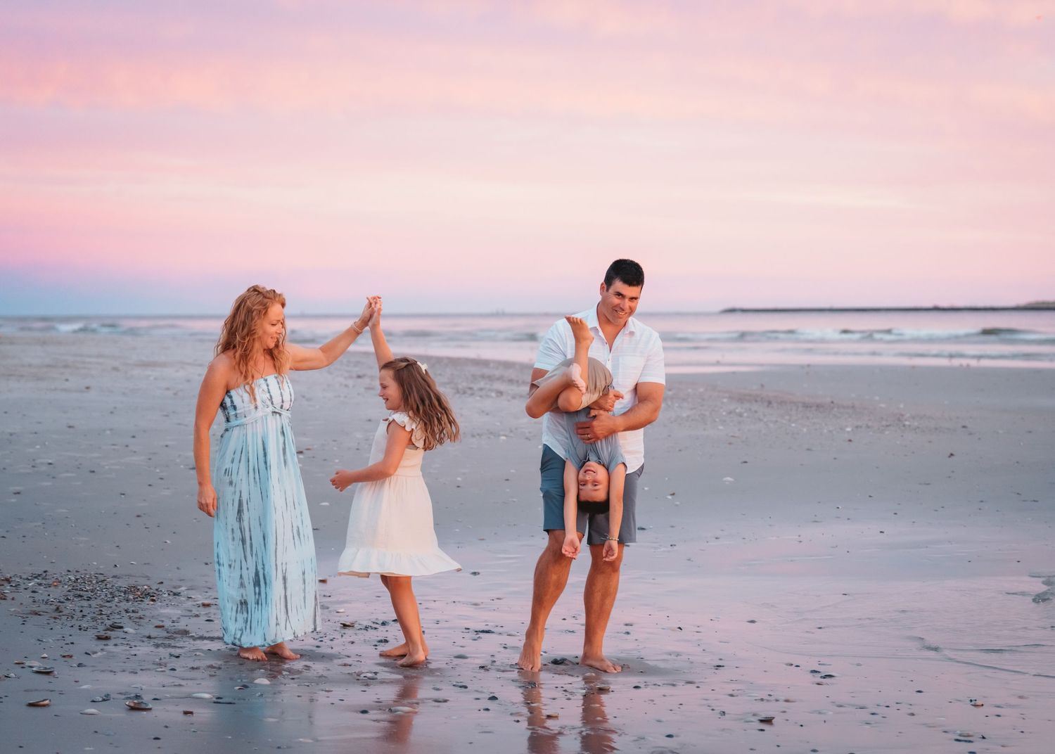 Family of four enjoying a playful moment on the beach at sunset with pastel pink and blue skies.