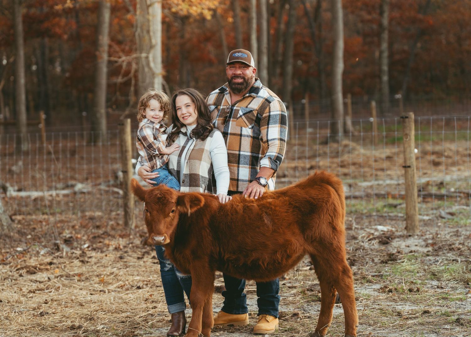Family of three posing with a brown highland calf in a fenced farm during autumn foliage season.