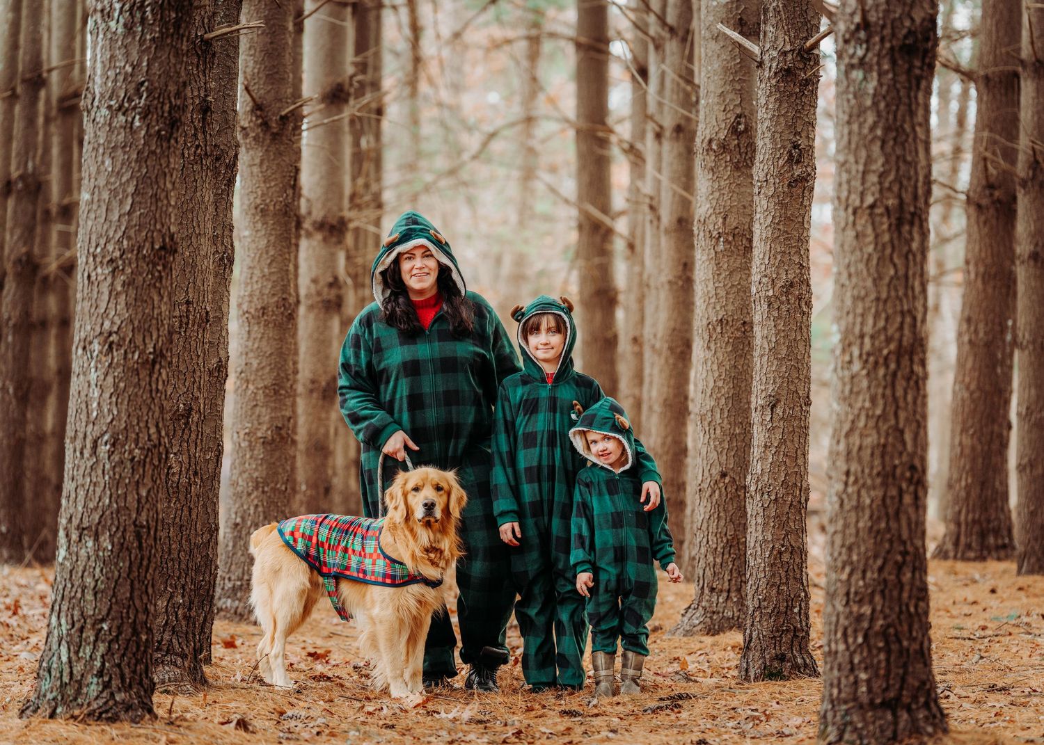 Mother and child in matching green plaid outfits walking with a golden retriever in pine forest.