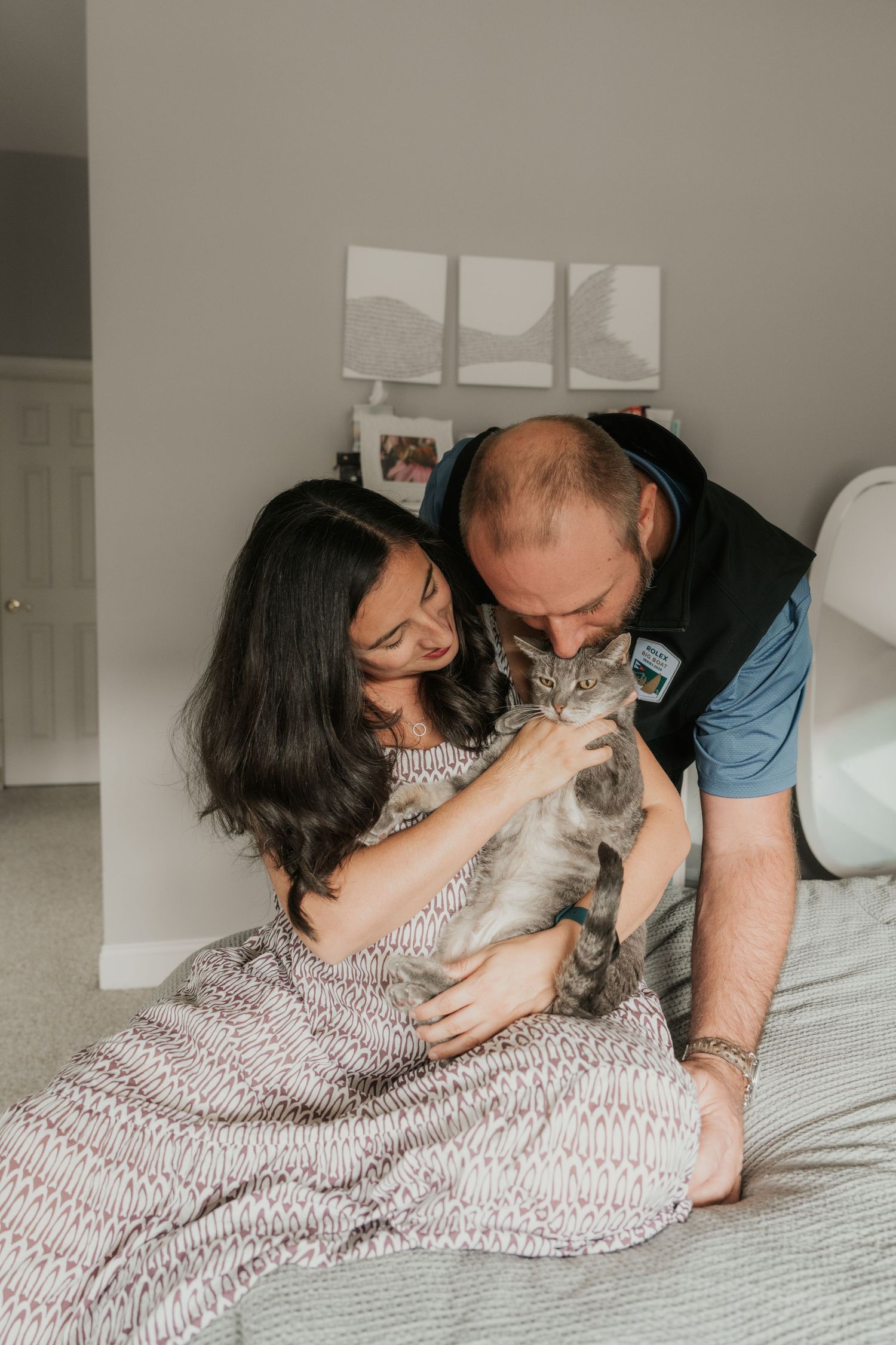 Couple sitting on a bed sharing a tender moment while petting their gray tabby cat indoors.