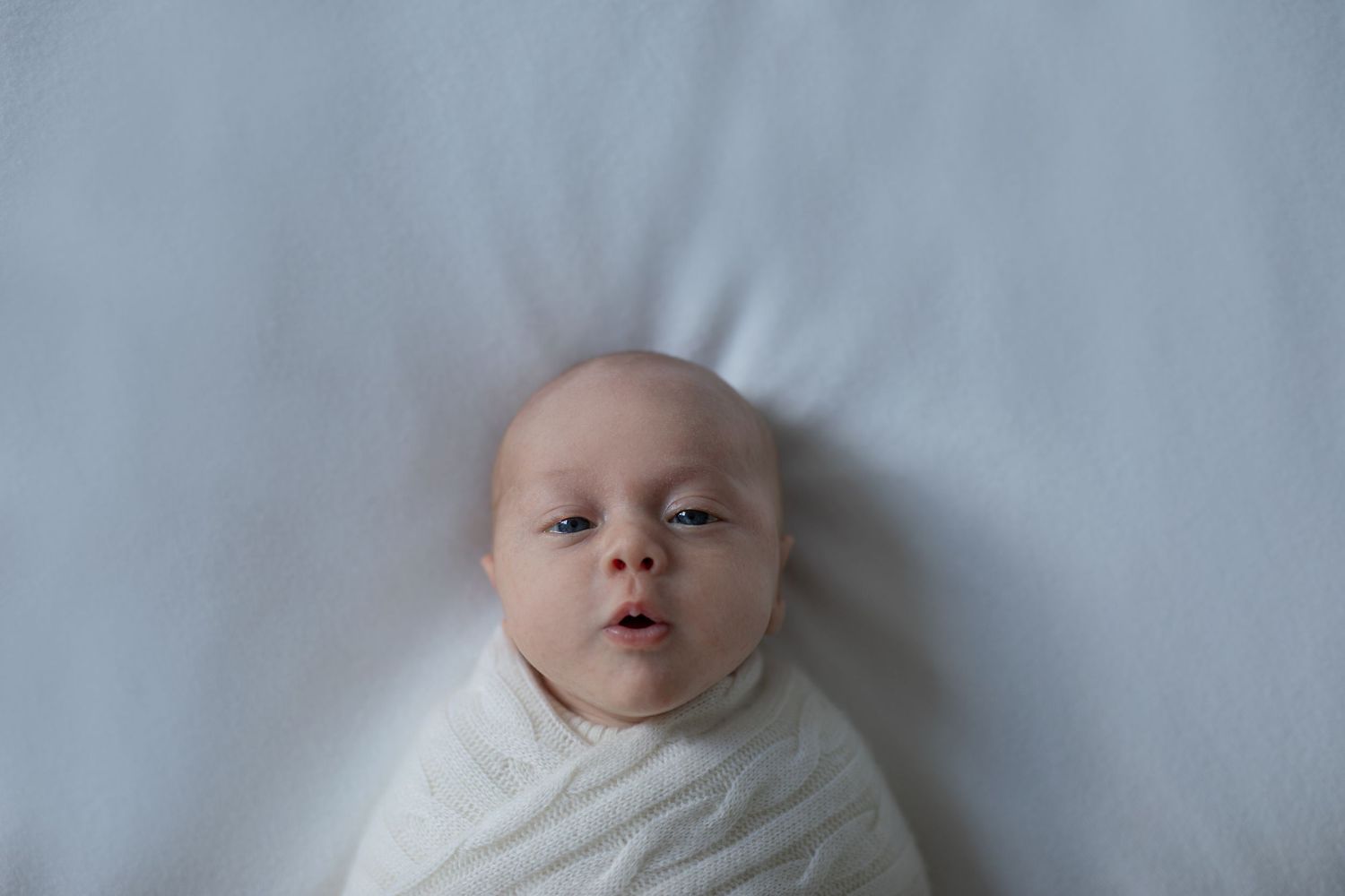 A sleeping infant wearing white lies on soft white bedding in a peaceful pose.