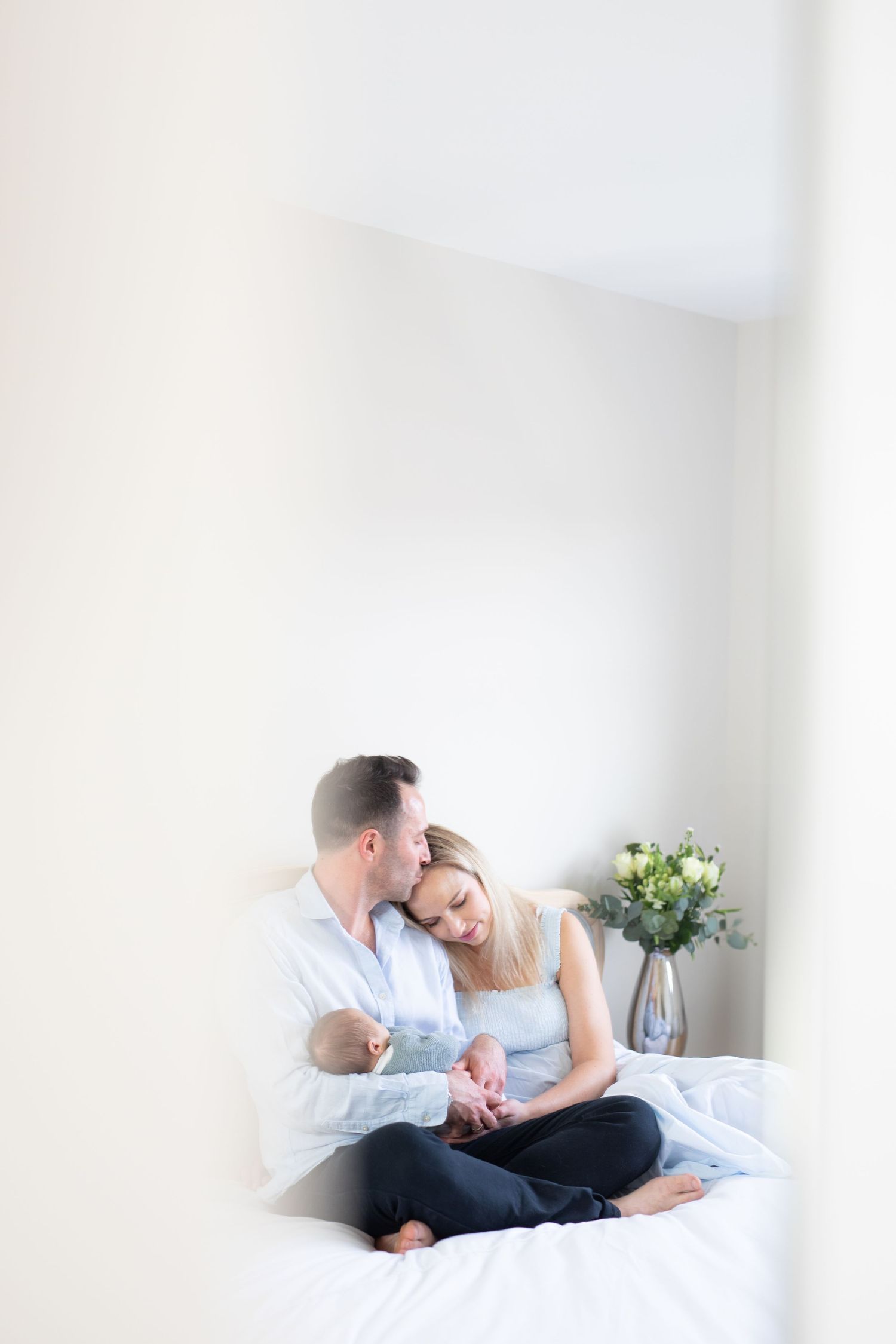 Intimate couple sharing a tender moment while sitting together on white bedding with flowers in vase.
