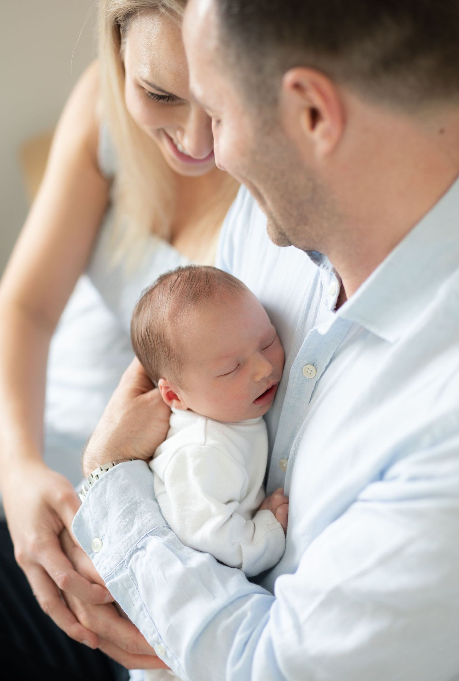 Parents holding and gazing lovingly at their sleeping newborn baby dressed in white clothing.