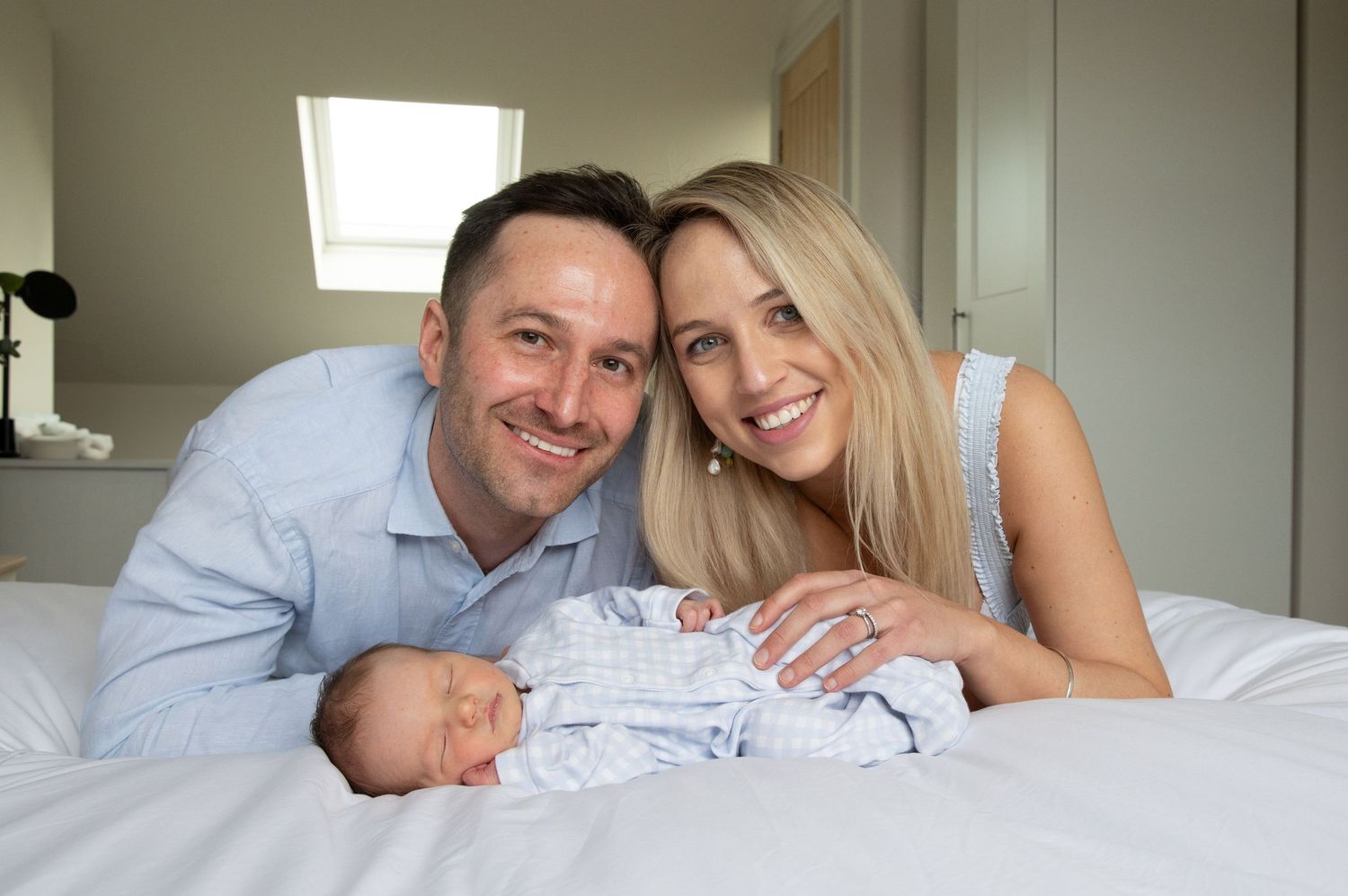 A couple lies on a white bed with their newborn baby wearing light blue clothing.