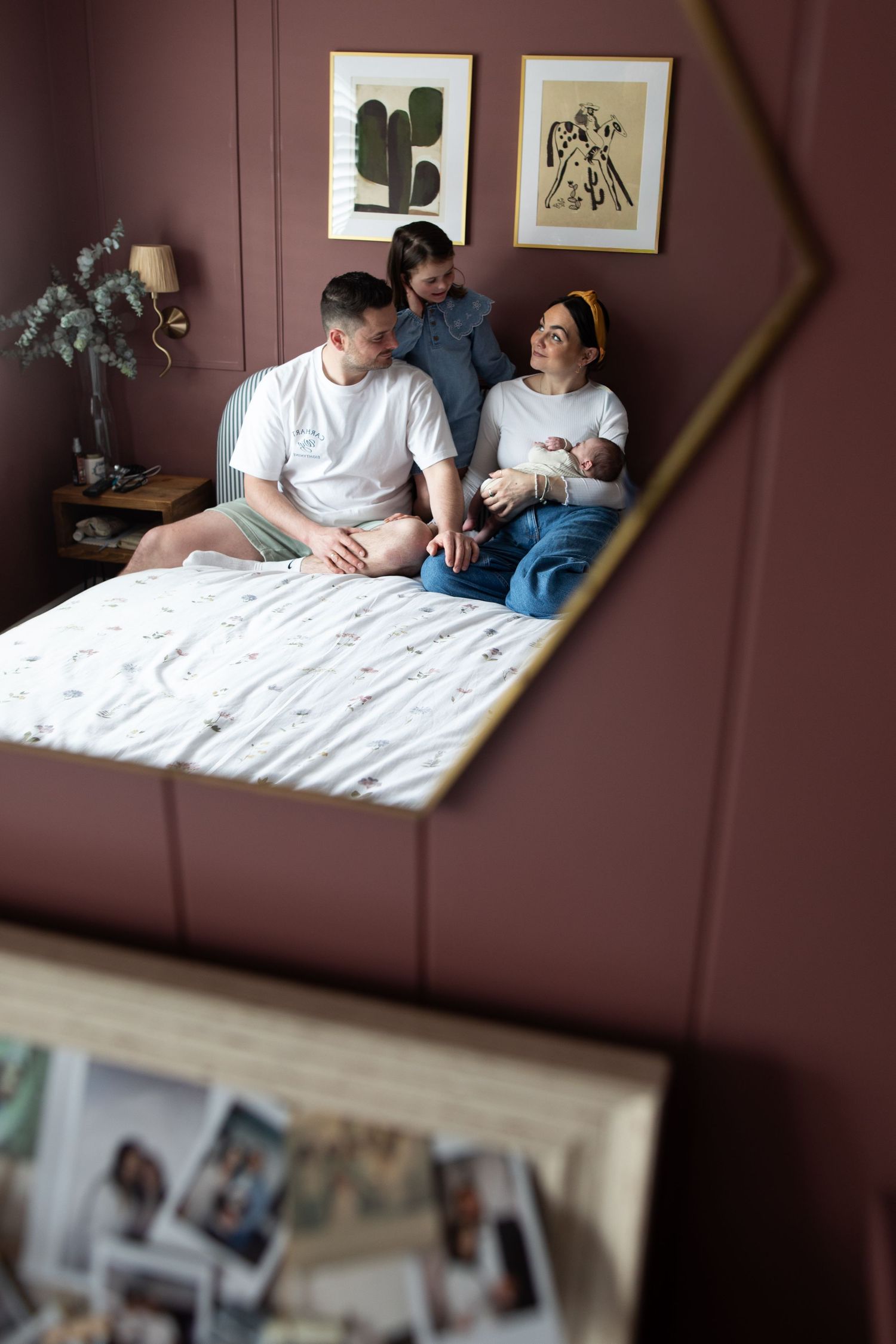 Family of three relaxing together on a bed in a cozy bedroom with brown walls and framed botanical art.