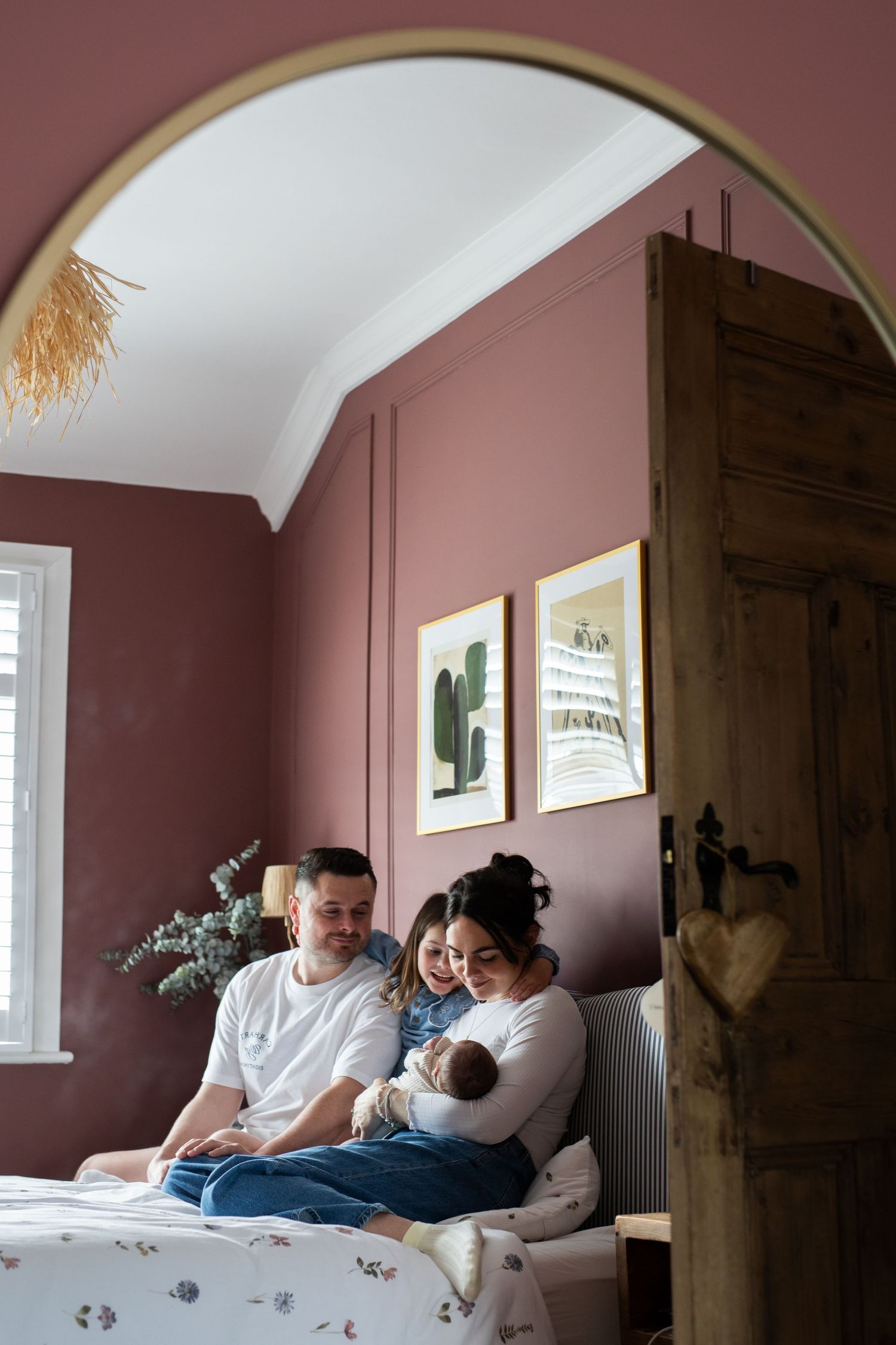 Family with newborn baby sitting on bed in pink bedroom, viewed through rustic wooden door and arched mirror.
