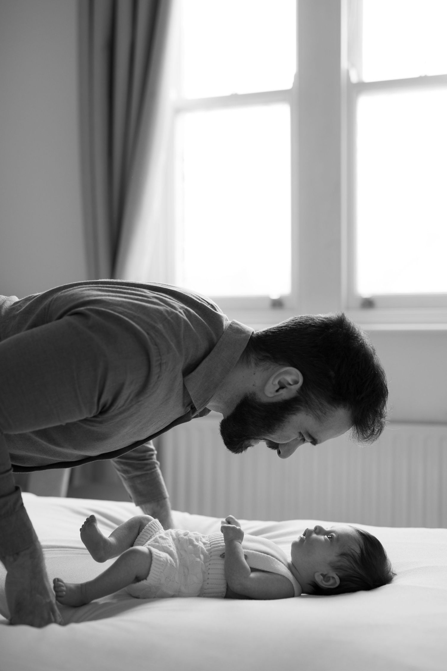 A tender black and white moment shared between a parent and baby lying on a bed near a bright window.