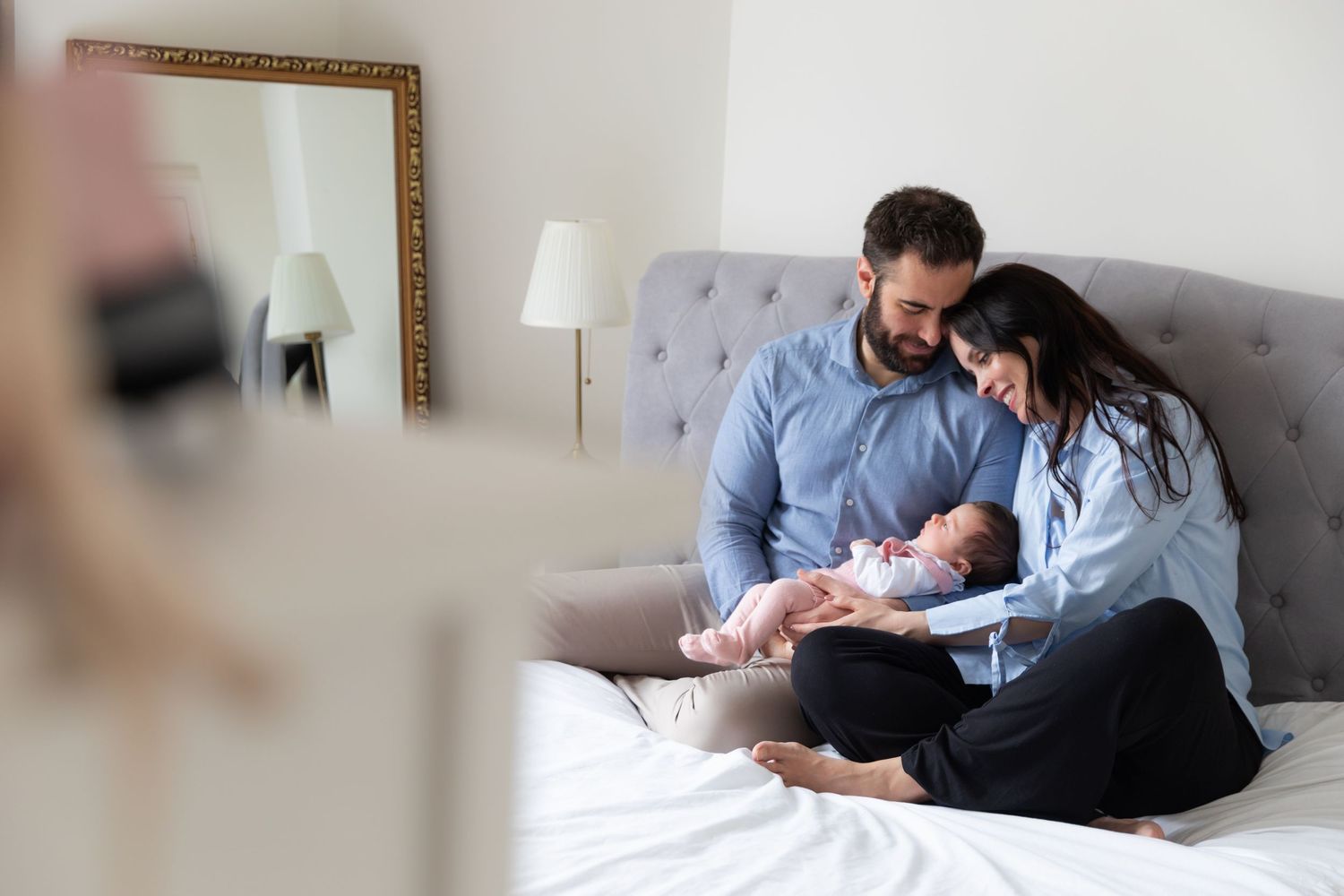 Parents sit together on a gray tufted bed, tenderly cradling their newborn baby while sharing an intimate family moment.