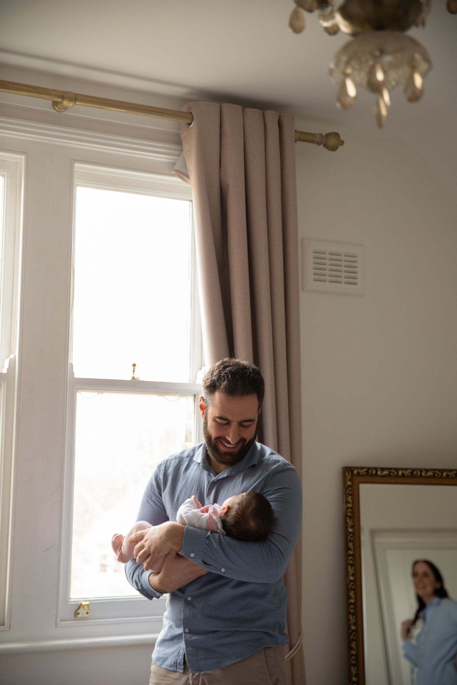 A parent in a light blue shirt stands by a bright window holding a tiny newborn baby in a cozy home setting.