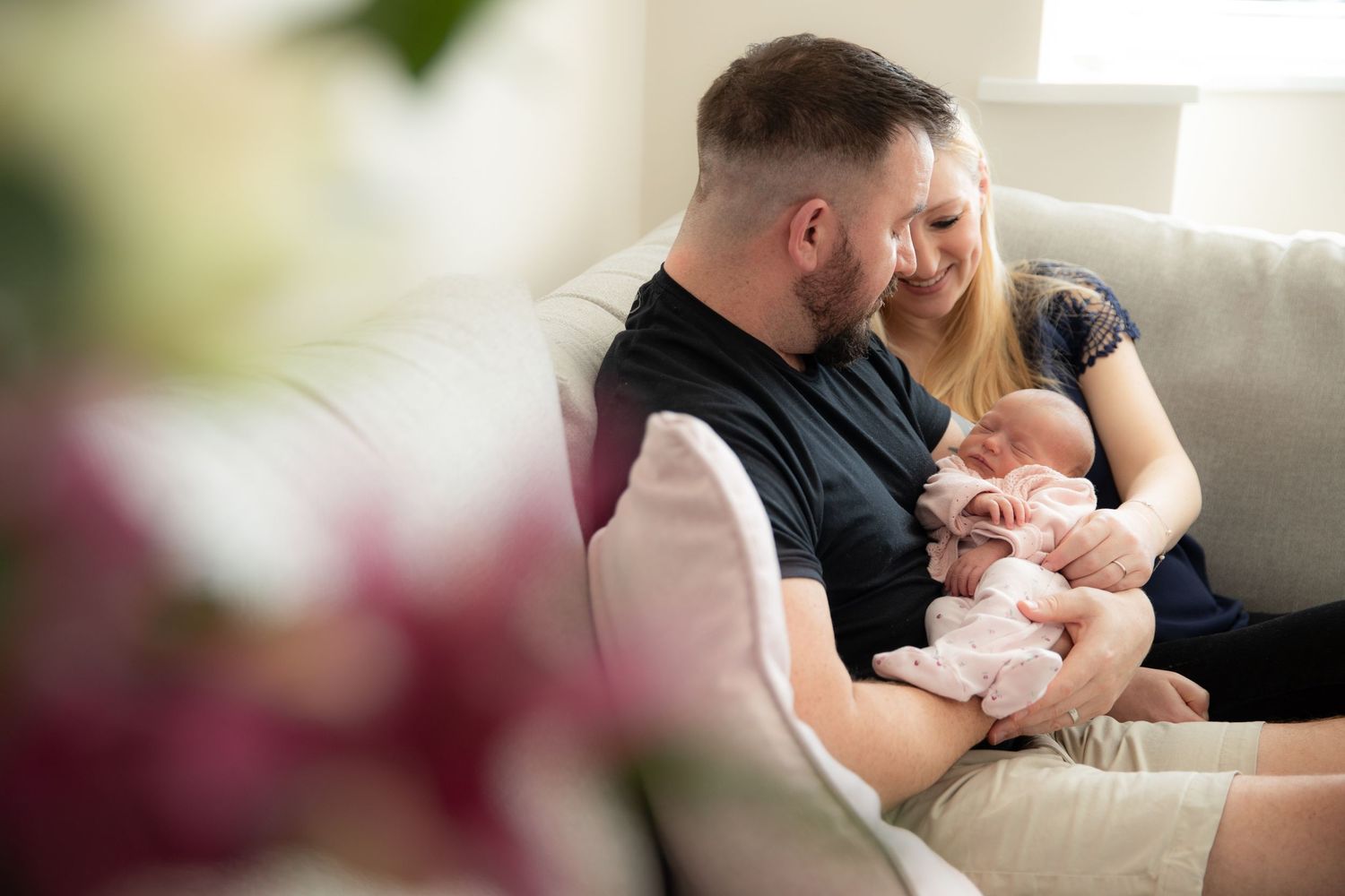 Parents sit together on couch tenderly holding and gazing at their newborn baby in sunlit living room.