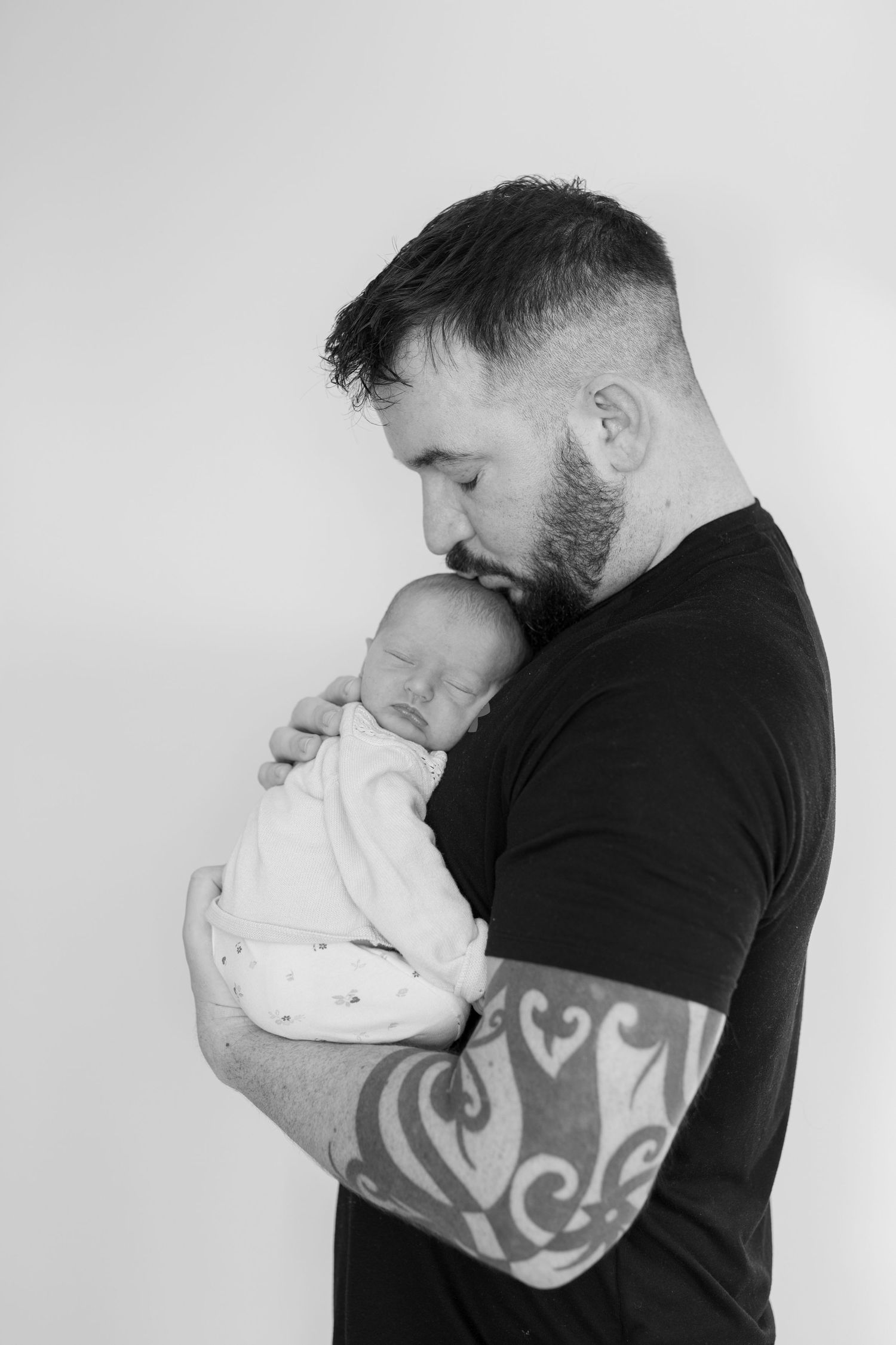 A tender black and white portrait of an adult in a black t-shirt with tribal tattoos holding a sleeping newborn baby.