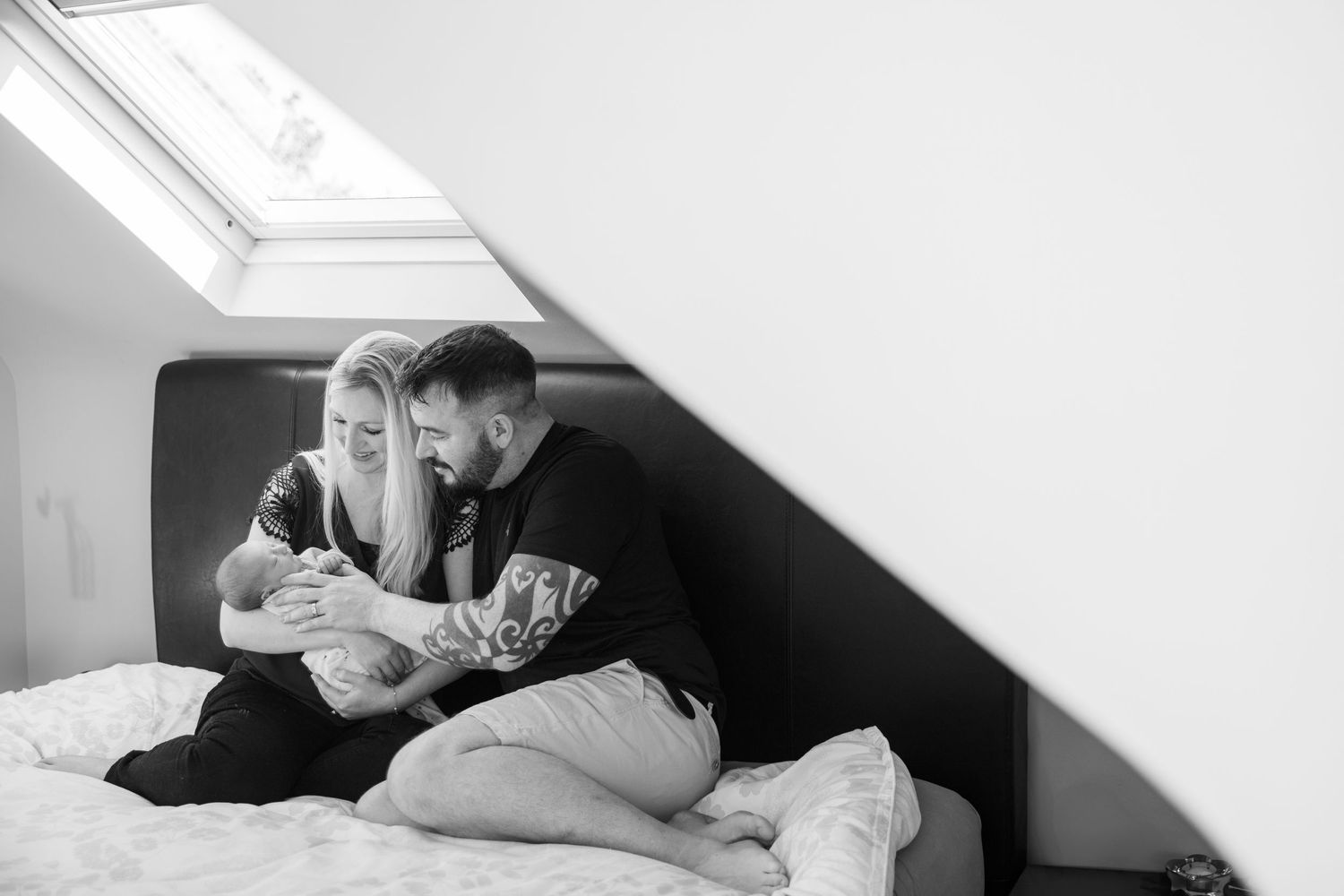 A couple sits together on a bed under a skylight window in an intimate black and white photograph.