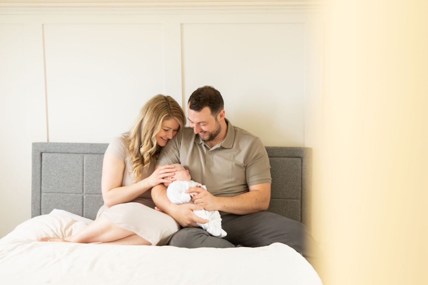 A family shares a tender moment together while sitting on a bed with grey headboard in natural lighting.