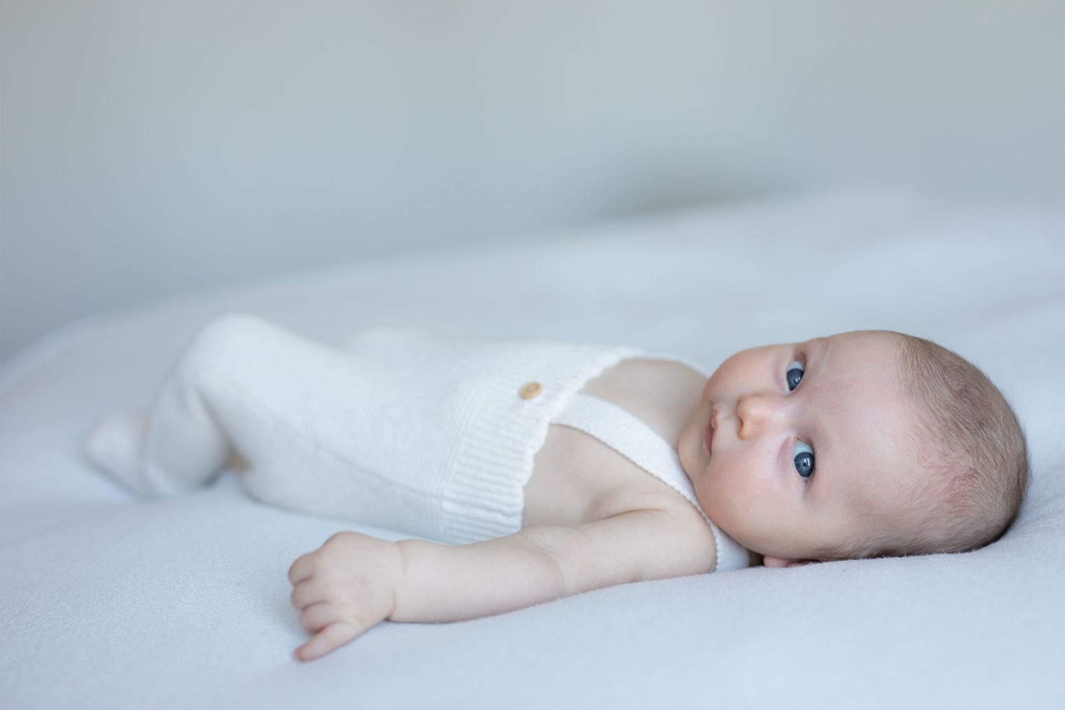 Infant in white clothes lying on light bedding with bright blue eyes looking up at the camera.