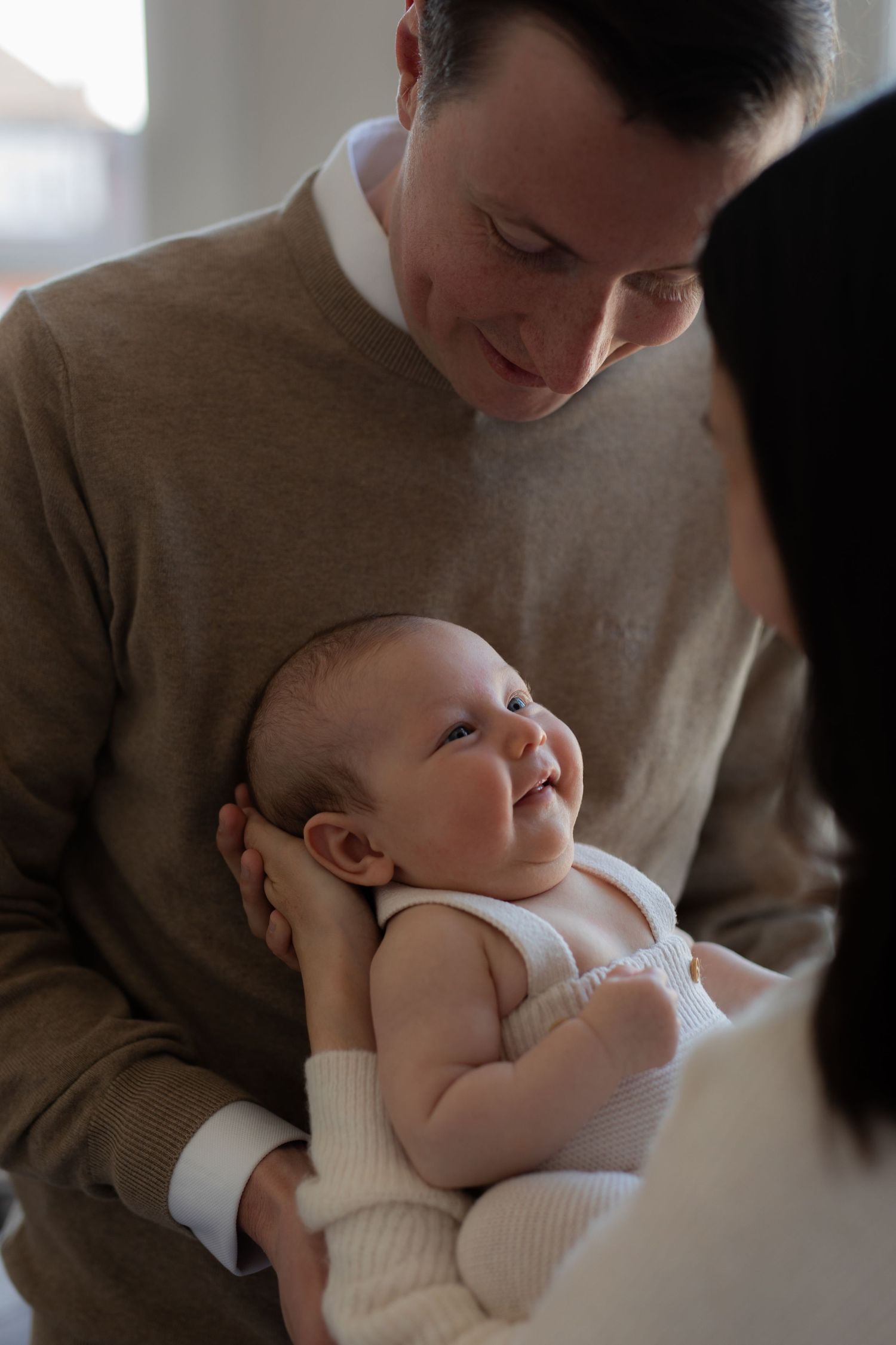 A tender moment with a baby in white clothing being cradled against someone wearing a tan sweater.