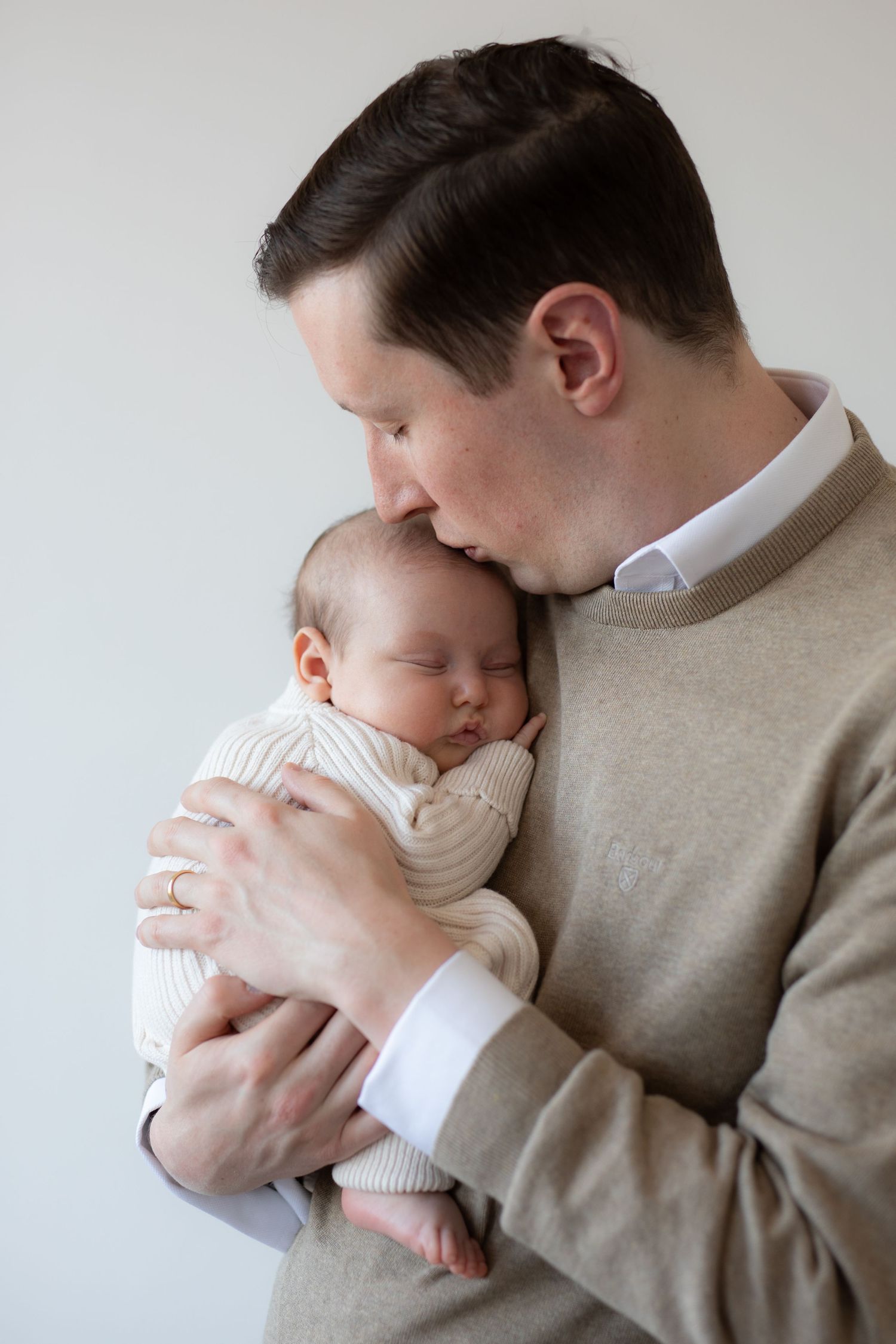 A tender moment as parent in beige sweater holds and kisses sleeping newborn baby wearing white knit outfit against gray wall.