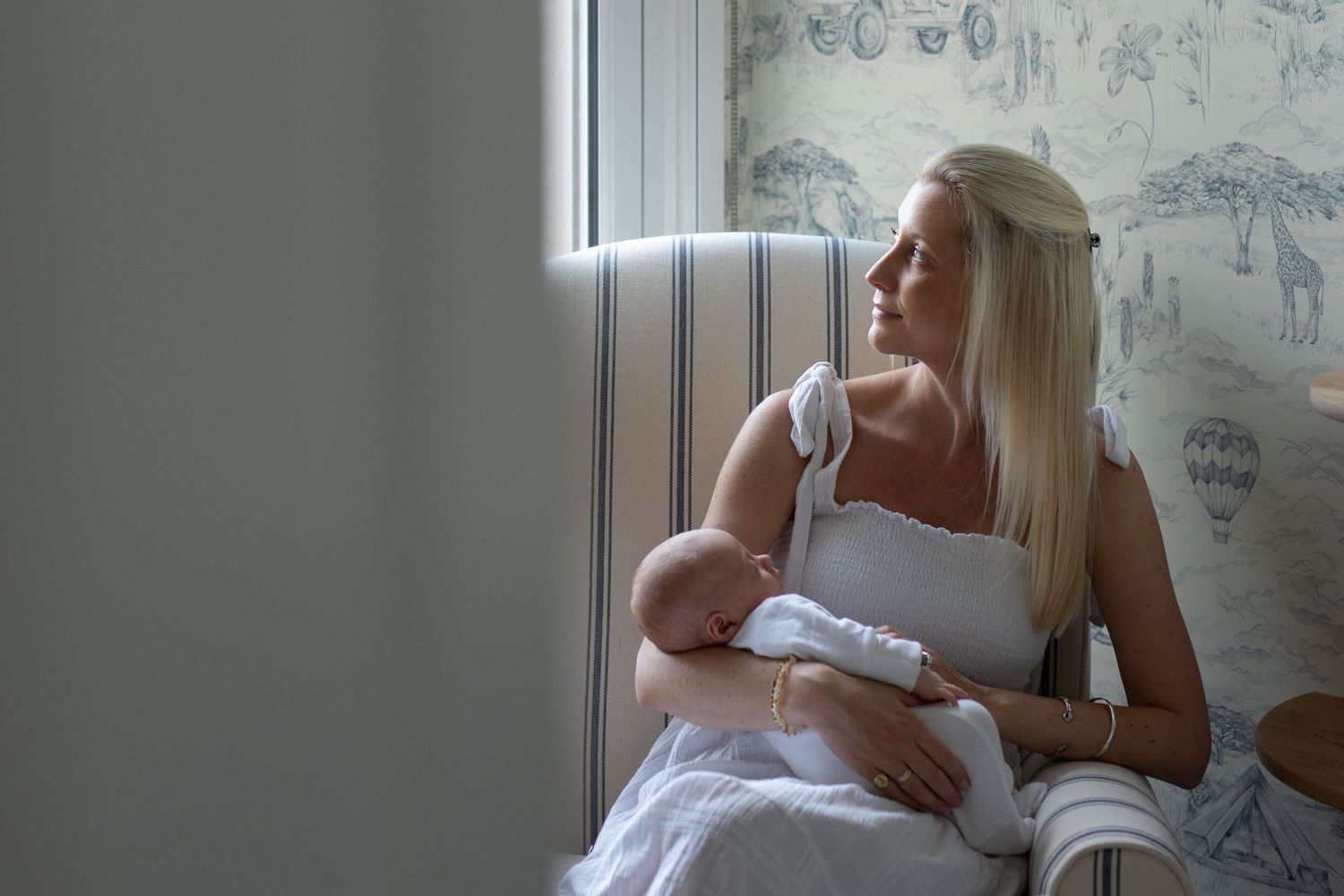 A parent in white clothing sits by a window while nursing a baby in a room with patterned wallpaper.