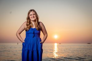A person in a vibrant blue dress stands by the ocean during sunset as golden light reflects on the calm water.
