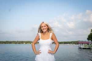 A person in a white dress poses joyfully by a scenic waterfront with boats and docks in the background.