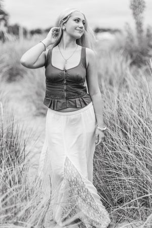 Black and white portrait of someone in a corset top and white skirt standing in tall grass field on a sunny day.