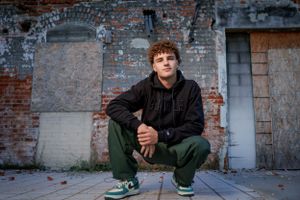 A person in dark clothing squats against an old brick wall backdrop in an urban setting.