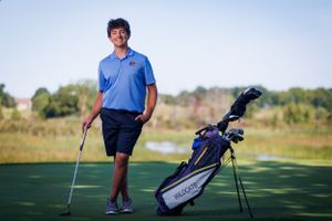 A golfer in blue polo and shorts stands on a golf course next to their golf bag during a sunny day.