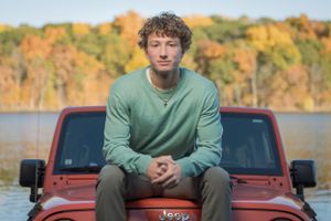 A person in a green sweater sits on the hood of a red Jeep with autumn foliage and water in the background.