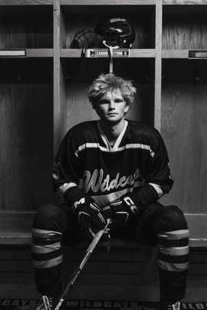 Black and white photograph of a hockey player seated in a locker room wearing a Wildcats jersey and gear.