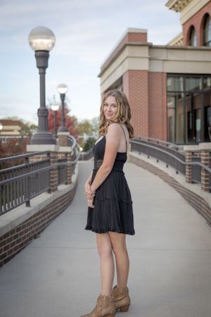 A figure in a black dress and brown boots poses on a brick-lined walkway with streetlamps and buildings in the background.