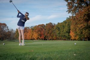 Golfer swings club on autumn golf course with vibrant orange and red foliage in background.