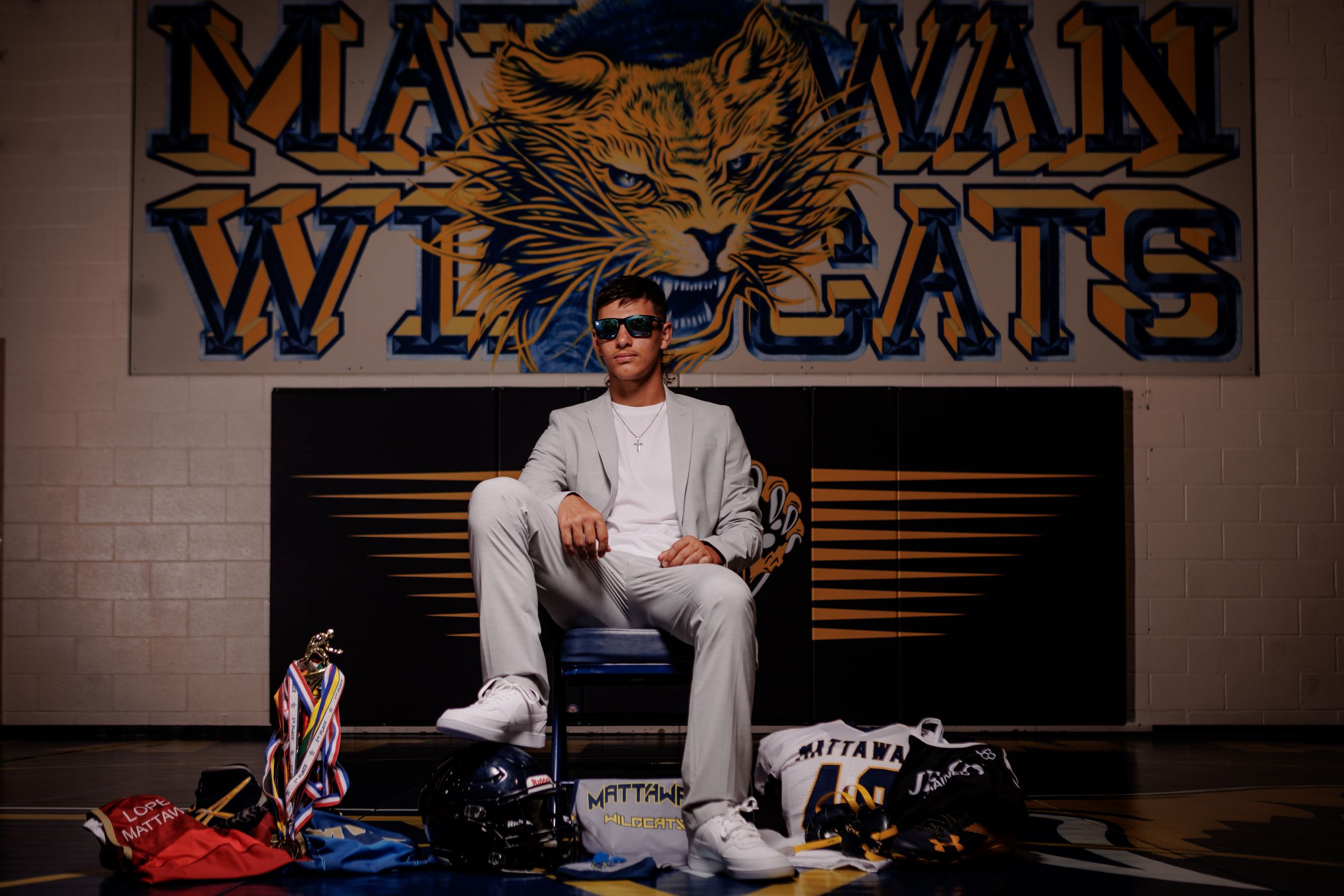 A coach sits on a bench under a Macewan Wildcats logo sign surrounded by athletic gear and equipment.