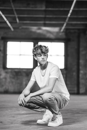 A black and white portrait of someone crouching in an indoor space wearing a white t-shirt, jeans and sneakers.