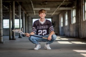 High school lacrosse player in jersey number 22 crouches in empty hallway holding lacrosse stick.