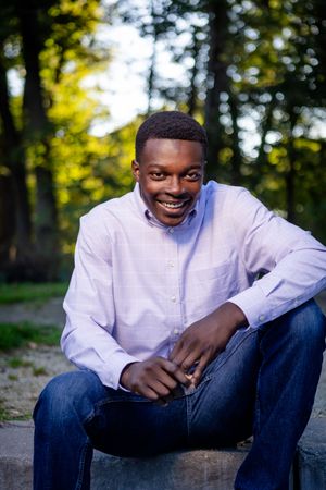 Outdoor portrait of someone in a light purple shirt and jeans sitting casually on a stone wall with sunlit trees in background.