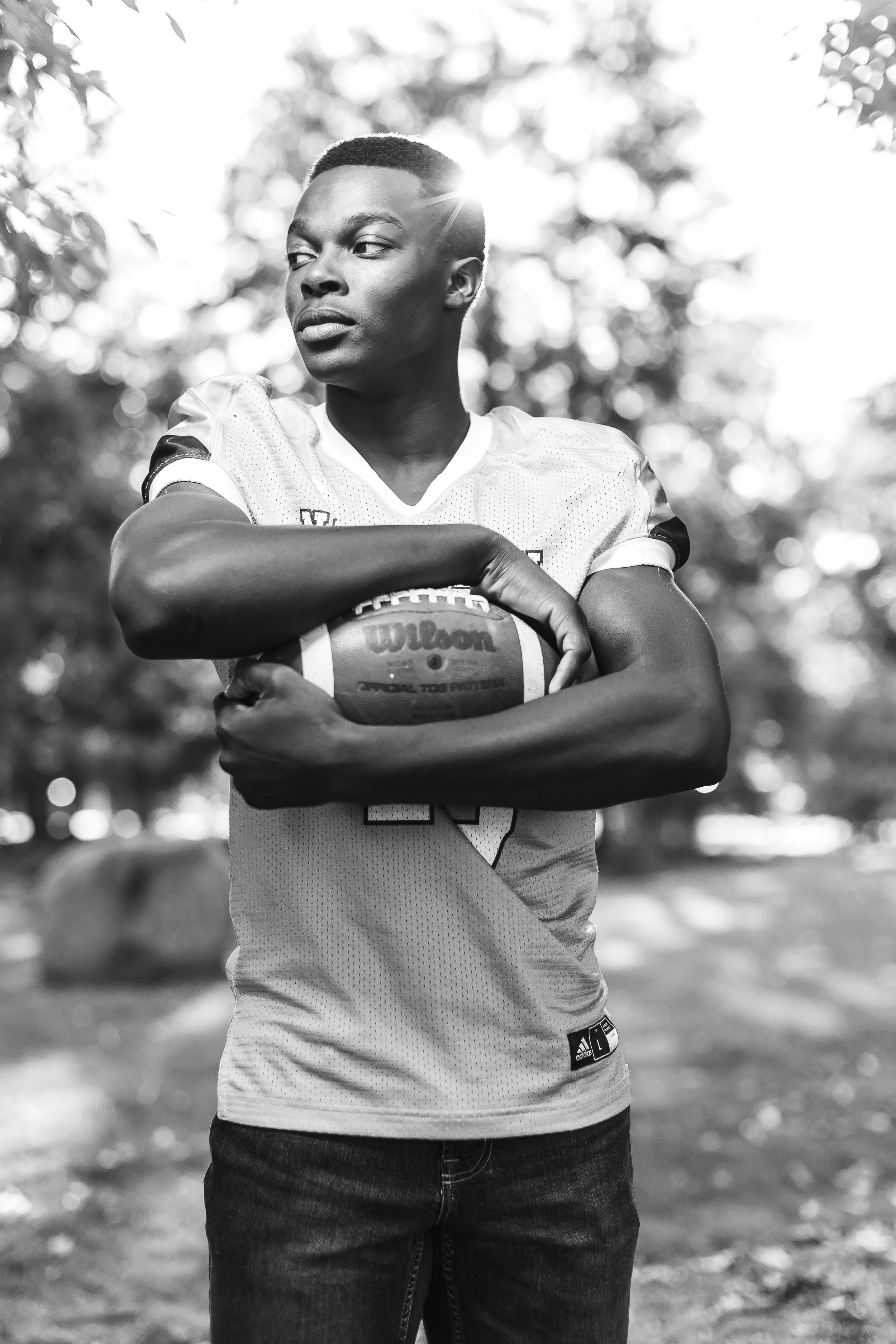 Black and white portrait of an athlete in casual attire holding a football outdoors with trees in background.