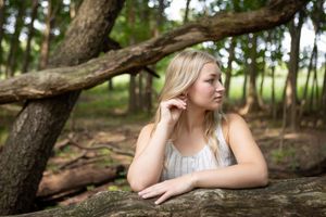 Someone poses thoughtfully in a natural forest setting with fallen logs and dappled sunlight.