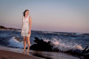 Someone in a white dress stands barefoot on a beach at sunset while waves crash against nearby rocks in the ocean.