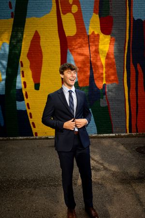 A person in a dark suit smiles and poses against a colorful graffiti wall with vibrant yellow and red patterns.