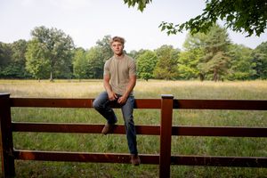 Someone sits casually on a wooden fence overlooking a peaceful green meadow with trees in the background.
