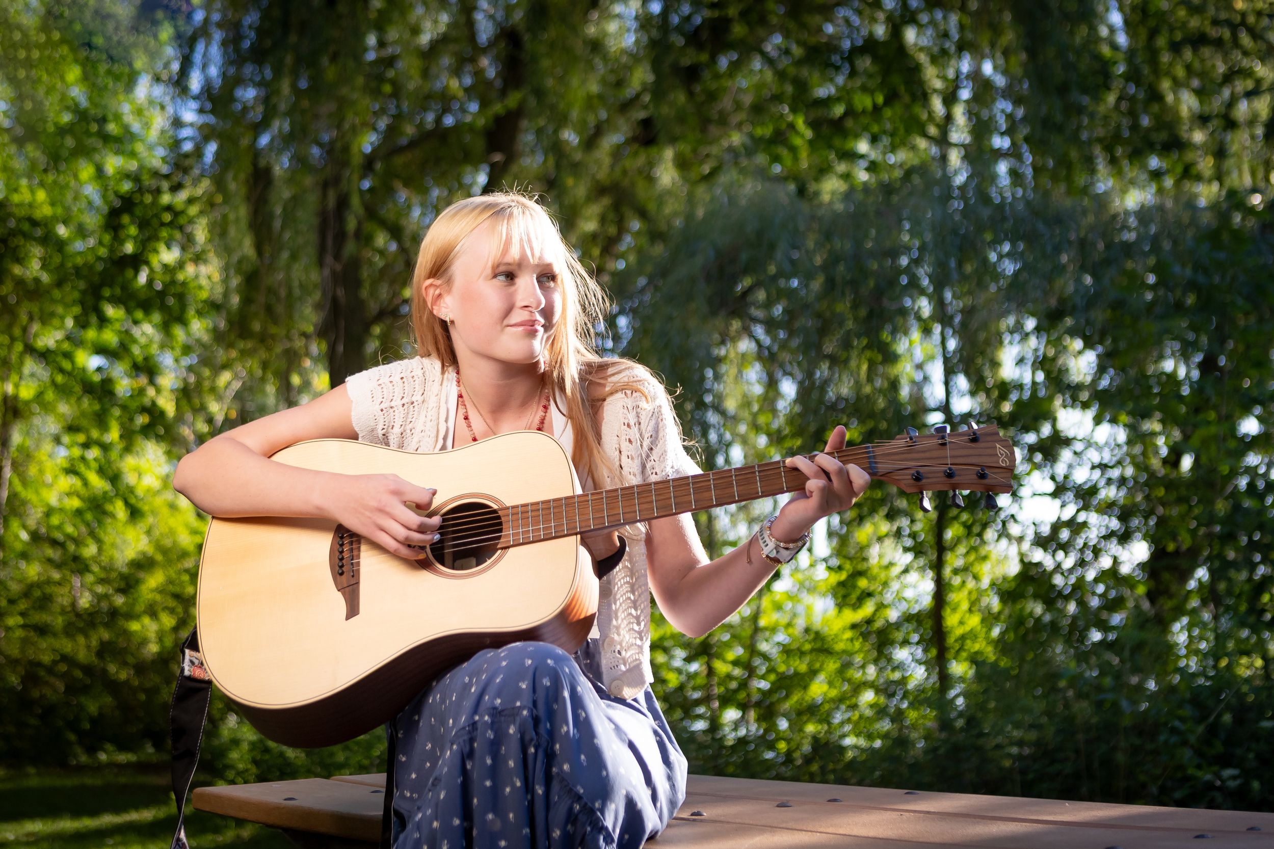 A musician plays an acoustic guitar outdoors in a natural wooded setting with sunlight filtering through trees.