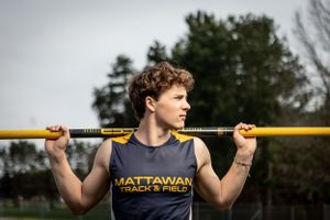 High school athlete wearing Mattawan track and field uniform holds pole vault bar across shoulders during outdoor practice.