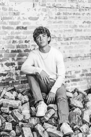Black and white portrait of person in white sweater sitting on pile of rocks near brick wall.