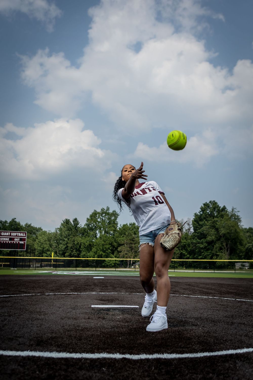 A softball player in uniform pitches a bright yellow ball on a dirt field under a cloudy blue sky.