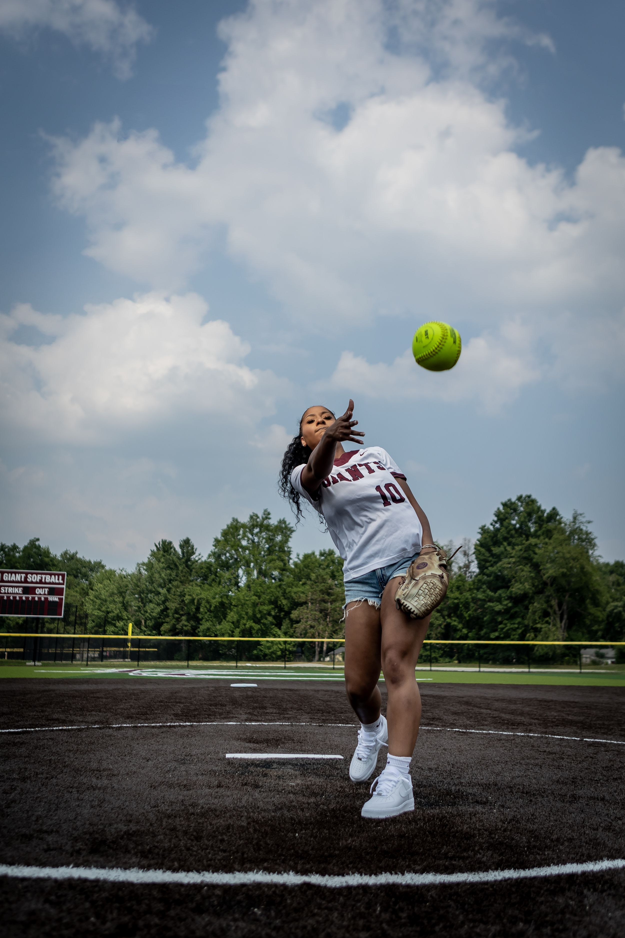 A softball player in uniform pitches a bright yellow ball on a dirt field under a cloudy blue sky.