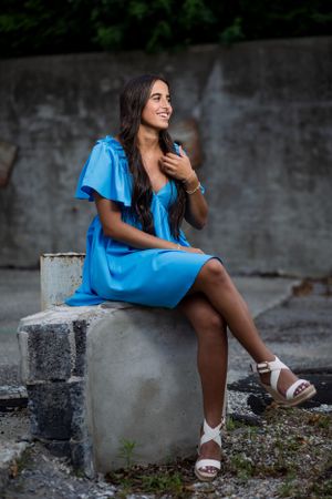 A person wearing a bright blue dress sits on a concrete structure outdoors, looking away while smiling.